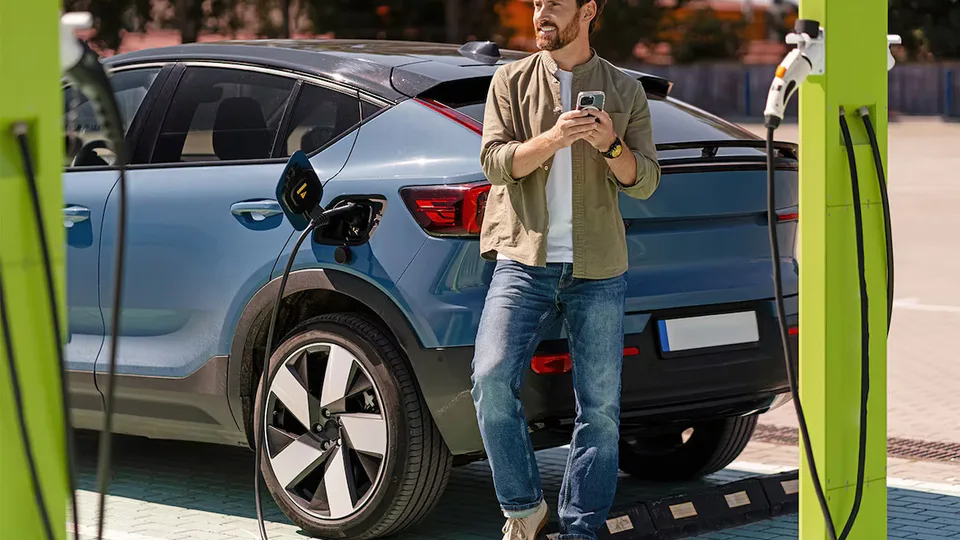 Man stands beside a blue electric SUV at a charging station, holding a phone while plugging in the charger.