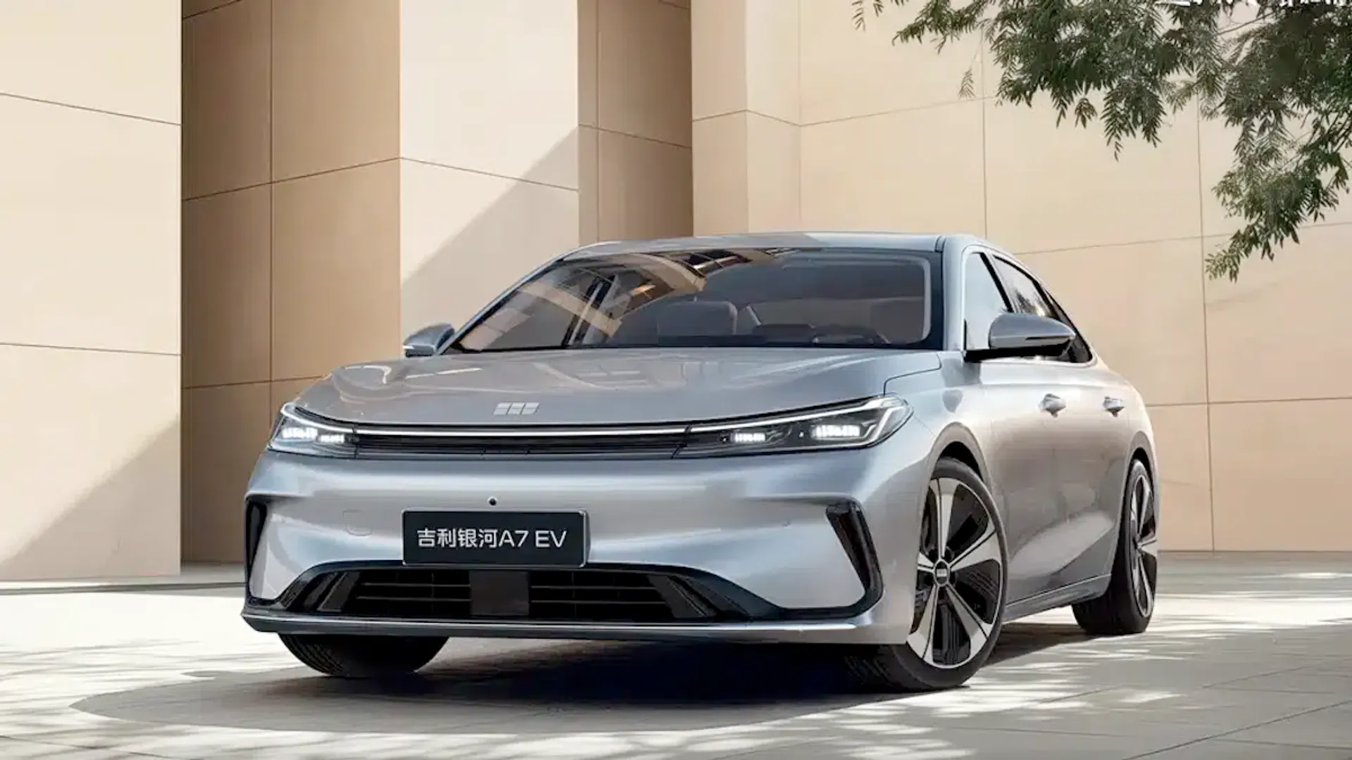 Silver Geely Galaxy A7 EV electric sedan shown in a three-quarter front view in front of a beige modern building, outdoor setting.
