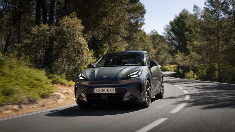 Front three-quarter view of a gray compact crossover driving on a winding forest road, sunlight filtering through trees, license plate 5836 NKX visible on the front bumper