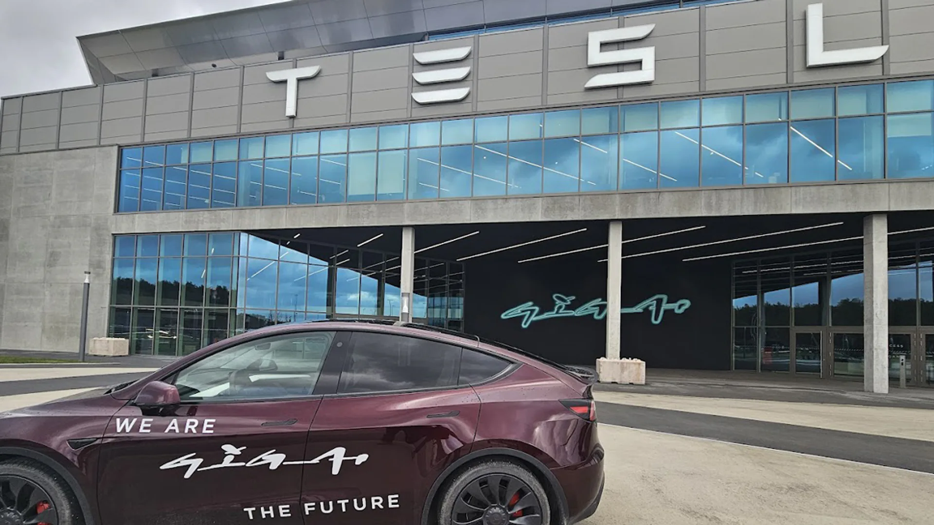 Maroon Tesla car with 'WE ARE GIGA' and 'THE FUTURE' decals parked outside a large glass-front Tesla showroom.01