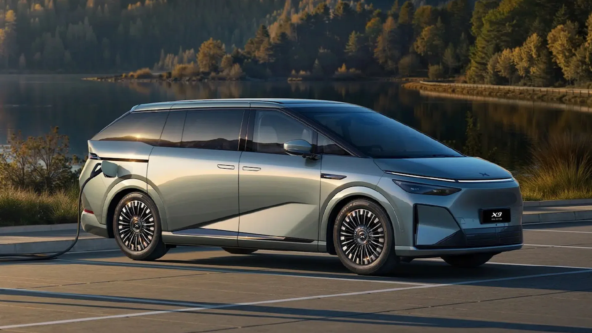 Silver electric minivan parked in a scenic lakeside lot, plugged into a charging cable with forested hills in the background.